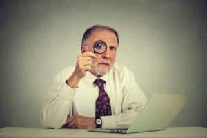 Serious business man in glasses skeptically looking through magnifying glass at you sitting at office table with laptop computer isolated gray background. Human face expression attitude, perception